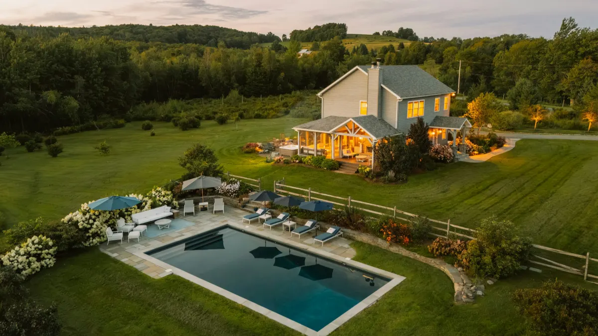 Grey timber house with a lit porch and a garden pool, seen from above at dusk