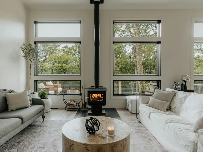 Living room with two pale sofas, a round stone coffee table and a black wood stove between tall windows