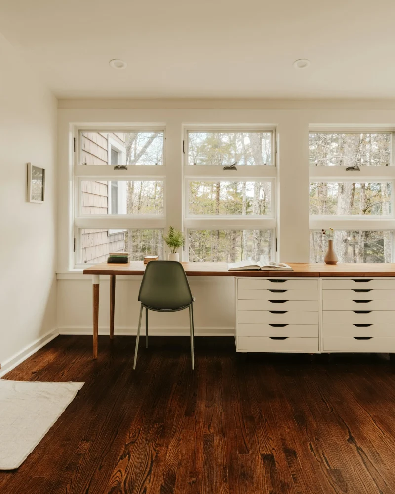 Studio with a long wooden desk, white drawers, two green chairs and a row of windows onto trees