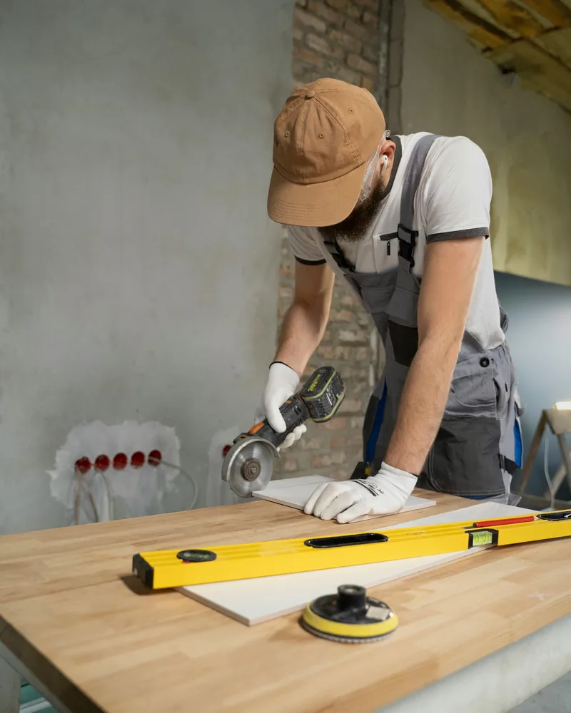 Tradesman in a cap and overalls cutting a wall tile with an angle grinder on a workbench