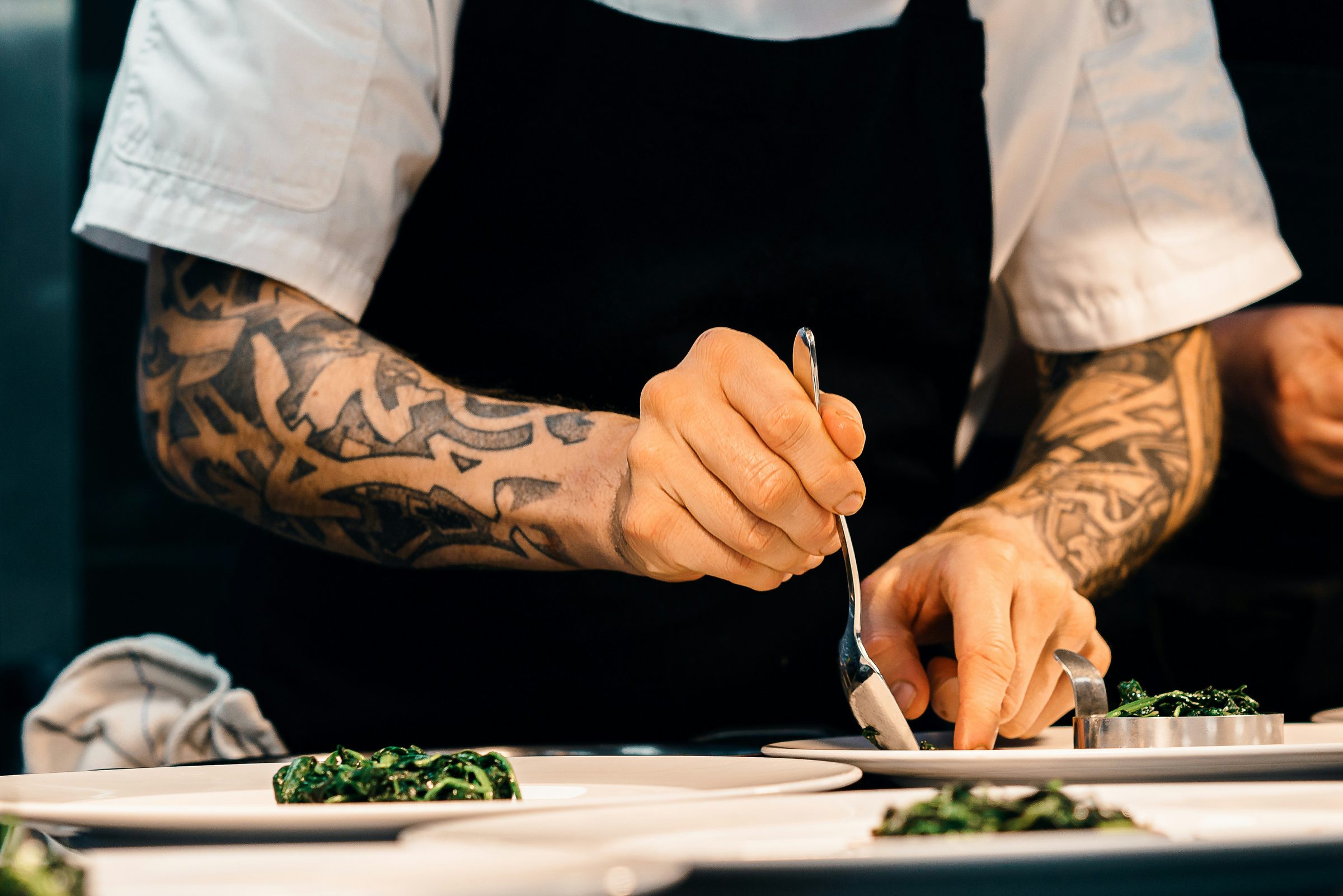 A chef plating fresh green vegetables on white plates at a restaurant kitchen pass